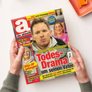 Titelblatt der Zeitschrift „die aktuelle“ mit großem roten Schriftzug „Todes-Drama um seinen Vater“ über dem Porträt eines Mannes in heller Jacke; weitere kleinere Porträts zeigen zwei lächelnde Frauen, dazu Textfelder mit Schlagzeilen wie „Blitz-OP gelungen!“ und „Irres Trennungs-Chaos“. Hände halten die Illustrierte vor hellorangem Tablett auf weißem Untergrund.