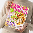 Eine nicht näher erkennbare Person in einem beigefarbenen Strickpullover hält ein schräg liegendes Kochmagazin mit dem Titel „essen & trinken“ und der Aufschrift „Aus Liebe zum Kochen“. Das Cover zeigt einen Salat und ein Gericht mit Hähnchen sowie die Texte „Saucen-Schule“, „Zeit für Tomaten“, „Workshop“, „Italien für Genießer“ und „Neue Hits mit Huhn“.