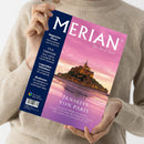 Helle Person in beigefarbenem Strickpullover hält eine Ausgabe des Reisemagazins „MERIAN – The Art of Travel“ mit violett-blauem Titelblatt. Abgebildet ist Mont-Saint-Michel bei Sonnenuntergang, reflektiert im Wasser. Sichtbare Textzeilen: „JENSEITS VON PARIS“, „Magischer Oman“, „Viva Valencia!“, „Legendäre Hotelbars“, „Bodensee“ sowie „Mit Google Maps Service“. Ausgabe 1/2025 mit weißem Barcode links unten.