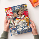 Titelblatt der Zeitschrift „Sport Bild“ mit rotem Logo links oben, Schlagzeilen wie „Bayern: Jagd auf WM-Stars“, „Kimmich exklusiv: So greifen wir den Titeln an“, „Klopp-Ärger in Leipzig“ und „Red Bull will Verstappen-Klausel abkaufen“. Abgebildet sind mehrere männliche Personen in Fußball- und Motorsportkleidung, eine goldene WM-Trophäe, farbiges Hintergrundlayout mit Blau, Rot und Gelb, gehalten von zwei Händen über hellem Untergrund.