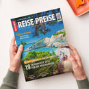 Magazin „Reise & Preise“ mit Titelbild eines sonnigen Küstenmotivs: türkisblaues Meer, weiße Felsklippen, grünes Laub und ein Strand mit Sonnenschirmen und Liegen. Sichtbare Texte: „Queensland – Australiens Naturwunder“, „Den Sommer verlängern – 19 traumhafte Ziele für die Nebensaison“, darunter „Istrien“, „Bangkok“, „Irland“ und „Marokko“. Gehalten von zwei Händen über heller Tischoberfläche mit orangefarbener Ablageschale.