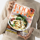 Magazin „Food and Travel Deutsch“ mit orangefarbener Schrift, gehalten von einer Person in beigem Pullover vor neutralem Hintergrund. Titelbild zeigt eine Schüssel asiatische Nudelsuppe mit Hühnerstreifen, Spinat, Chili und Essstäbchen. Sichtbare Texte: „Rezepte für den Frühling“, „Irisch backen leicht gemacht“, „Neue Safari-Lodges“, „Stadthotels unter 200 €“, „Malaysias Gourmetinsel Langkawi“, „Kulinarik-Hotspot Cadiz“, „Cordoba“, „Skifahren im Spätwinter“.