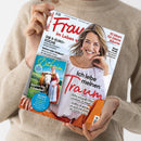 Beige gekleidete Person hält schräg eine Zeitschrift mit weißem, glänzendem Cover und dem Titel „Frau im Leben“; darauf eine lächelnde Frau im weißen Oberteil. Sichtbare Texte: „Die 5-Euro-Küche“, „Gehen oder bleiben?“, „10 Ideen für kleine Räume“, „Neue Mode“, „Ich liebe meinen Garten“ sowie „Bayern für Entdecker“ und „Bayern von A–Z“.