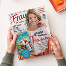 Auf einer weißen Fläche halten zwei Hände die schräg liegende Zeitschrift „Frau im Leben“ mit weiß-rotem Cover; darauf eine blonde Frau in weißer Bluse sowie die Texte „Ich lebe meinen Traum“, „10 Ideen für kleine Räume“, „Neue Mode“, „Die 5-Euro-Küche“ und „Bayern für Entdecker“. Oben rechts liegt teilweise ein orangefarbenes Notizbuch mit Verschluss.