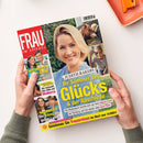 Auf weißer Fläche hält eine Hand ein schräg liegendes Hochglanzmagazin „FRAU IM SPIEGEL“ mit rotem Titelbalken. Das Cover zeigt Judith Rakers, die Schlagzeile „Ihr Sommer des Glücks“ sowie kleinere Beiträge zu Thomas Müller, Christine Neubauer, Kate & Louis und einem Pferd; oben rechts liegt ein orangefarbenes rechteckiges Objekt.