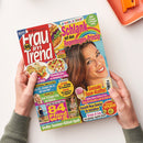 Buntes, schräg gehaltenes Magazin „frau im trend“ auf weißer Fläche, von zwei Händen mit grauen Ärmeln gehalten; auf dem Cover ein weibliches Model, Gemüsegerichte, Kuchen und Eis. Sichtbare Texte: „Je bunter, je besser!“, „Schlank mit dem Regenbogen-Prinzip“, „Großer Sommer-Rätsel-Spaß“, „84 Pfund weg“ und „Geniale Spar-Kniffe“; oben rechts liegt ein orangefarbenes Bürogerät.