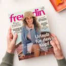 Schräg auf einem weißen Tisch liegende Zeitschrift „freundin“, von zwei Händen mit grauen Strickärmeln gehalten; das Cover zeigt eine weibliche Person mit Strohhut, blauem Strickpullover und weißer Bluse. Sichtbare Texte: „10 gute Neuigkeiten für unsere Gesundheit“, „Neue Rezepte mit Tomaten“, „Was hilft gegen den Hass?“, „Urlaub für den Kopf“ und „Sommer-schön!“. Oben rechts liegt ein orangefarbenes Tablett.