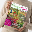 Eine Person im beigefarbenen Strickpullover hält eine schräg fotografierte Zeitschrift mit grün-pinkem Titel „GartenFlora“ und der Aufschrift „September 2026“. Das Cover zeigt eine blühende Gartenfläche mit roten, pinken und weißen Blumen sowie ein kleines Foto einer Frau im Garten; weitere Texte lauten „gärtnern, erleben und genießen“, „So werden Sommerblumen gezüchtet“, „Unkomplizierte Mini-Kiwis für den Garten“ und „Magischer Spätsommer in Burgunderrot“.