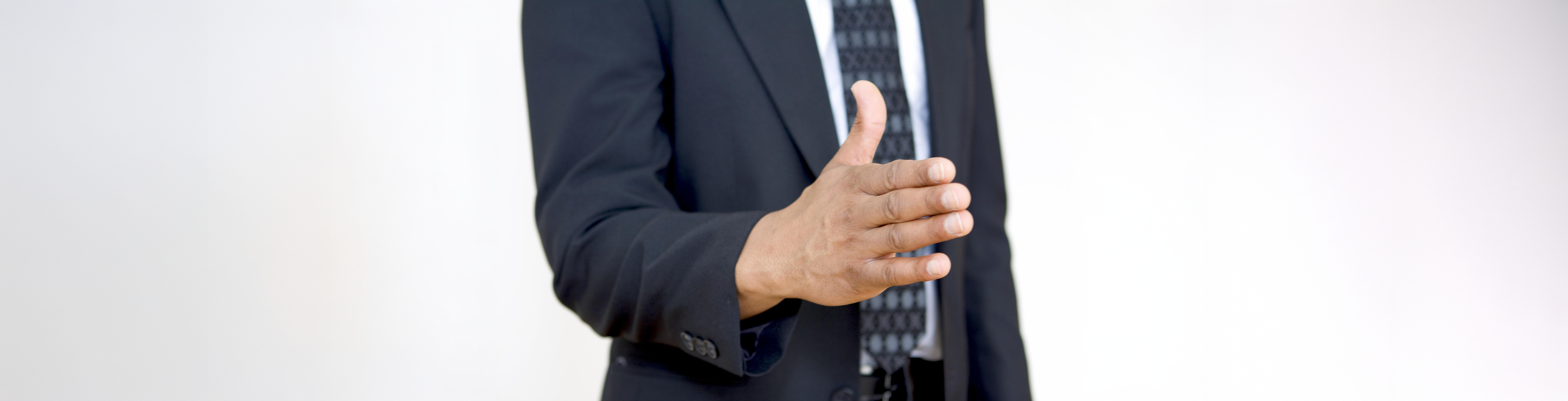 Männliche Person im schwarzen Anzug mit weißem Hemd und gemusterter Krawatte, frontal fotografiert, reicht mit ausgestrecktem rechten Arm und geöffneter Hand zum Händedruck; Bildausschnitt zeigt nur Oberkörper bis zur Hüfte, vor neutralem, hellgrauem Hintergrund.