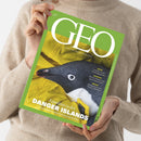 Grün gerahmtes GEO-Magazin mit dem Titel „GEO“ und den Aufschriften „AUGUST 2026“ sowie „DIE WELT MIT ANDEREN AUGEN SEHEN“, gehalten von einer Person im beigen Strickpullover. Das Cover zeigt einen Pinguin in gelber Kleidung; darunter steht „Expedition zu den DANGER ISLANDS – Deutschlands weißer Fleck am Ende der Welt“, daneben Themen zu Käfern, Sokrates und Mexiko.