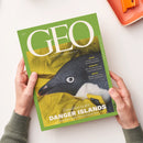 Grünes GEO-Magazin „AUGUST 2026“ mit großem weißem GEO-Schriftzug, dem Untertitel „DIE WELT MIT ANDEREN AUGEN SEHEN“ und dem Titel „Expedition zu den DANGER ISLANDS – Deutschlands weißer Fleck am Ende der Welt“. Das Cover zeigt einen schwarz-weißen Pinguin in gelber Kleidung; zwei Hände halten das schräg liegende Magazin vor hellem Hintergrund.