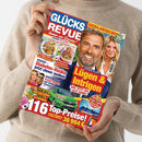 Hochformatige Zeitschrift „GLÜCKS REVUE“ mit rotem und blauem Cover, gehalten von einer Person ohne sichtbares Gesicht in beigefarbenem Strickpullover. Sichtbare Texte: „EXTRA-RÄTSELHEFT“, „35x Ratespaß“, „Lecker! Die besten Rezepte zum Schlemmen“, „Diabetes-Risiko jetzt clever senken“ sowie „Lügen & Intrigen“; abgebildet sind mehrere Personen, Speisen, ein grünes Auto und ein Fernseher.