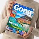 Eine Person im beigefarbenen Strickpullover hält schräg ein Magazin „Gong“ mit blauem Titelbild und der Aufschrift „Ihr TV-Programm vom 22.8. bis 28.8.2026“. Das Cover zeigt eine Küstenstadt auf Sizilien sowie die Texte „Inseln zum Verlieben“, „Zauberhafte Reisetipps fürs Mittelmeer“, „Der große Fußball-Bundesliga-Planer 2026/27“, „Raffinierte Fisch-Rezepte“ und „Ein bewegtes Leben“.