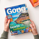 Auf weißer Fläche hält eine Person mit zwei Händen ein schräg liegendes Magazin mit blauem Titelbild „Gong“ und der Aufschrift „IHR TV-PROGRAMM VOM 22.8. BIS 28.8.2026“; abgebildet sind die Küstenstadt Cefalù, Meer und Felsen. Sichtbare Texte: „Inseln zum Verlieben“, „Zauberhafte Reisetipps fürs Mittelmeer“, „Bundesliga Planer“, „Frisch auf den Tisch“ und „Ein bewegtes Leben“; oben rechts liegt ein orangefarbenes Objekt.