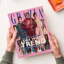 Rosa GRAZIA-Modemagazin mit drei Personen auf dem Cover: einem Mann links sowie zwei Frauen in der Mitte und rechts, gekleidet in Denim, roter Lederjacke und rosafarbenem Kleid. Sichtbare Texte: „DIE GROSSE RUNWAY-EDITION“, „DAS WIRD TREND“, „LEDER-STYLES“, „DOUBLE-DENIM“, „NEW-WHITE“ und „IM SPOTLIGHT: Charlize Theron + Sienna Miller + North West + Blake Lively“; das Magazin wird von zwei Händen gehalten, rechts oben liegt ein orangefarbenes Objekt.