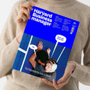 Person im cremefarbenen Strickpullover hält schräg eine Zeitschrift mit blauem Cover. Lesbare Texte: „Harvard Business manager“, „Das Wissen der Besten“, „Wie Game Boy und Crocs zu Ikonen wurden“, „Warum Chatbots Ikea umgarnt“, „Zahlen sich immersive Erlebnisse aus?“, „Wann es Zeit ist, Prioritäten neu zu setzen“, „Verhandeln am Limit“, „Nö.“ und „Jetzt gib schon auf…“.