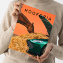 Weibliche Person im beigefarbenen Strickpullover hält ein schräg ausgerichtetes, orangefarbenes Magazin mit dem Titel „HOOPRIA“ und der Aufschrift „DIE KUNST DER PAUSE“. Das Cover zeigt ein braunes Känguru, einen grünen Oldtimer und helles Stroh; Hände und Unterarme sind sichtbar, das Gesicht ist nicht abgebildet.
