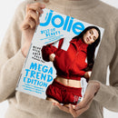 Diagonal gehaltenes Magazin „jolie“ mit weißem Cover und türkisblauer Schrift, in den Händen einer Person mit beigefarbenem Strickpullover. Abgebildet ist eine Frau mit langem schwarzem Haar in roter Jacke und roter Hose; sichtbare Texte: „BEST OF BEAUTY“, „MEGA TREND EDITION“, „MEHR GELD ABER EASY“, „10 geniale Tipps“, „New-Season-Make-up“, „Die neuen Blond-Töne“, „Düfte, die nicht jeder hat“, „Leder“, „Seventies“, „Denim“, „Strick“, „Layering“ und „Sporty“.