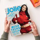 Aufgeschlagene Modezeitschrift „jolie“ in den Händen einer Person; auf dem Cover eine Frau mit langen dunklen Haaren in roter Jacke und roter Hose, darüber große blaue Schrift „MEGA TREND EDITION“ und „BEST OF BEAUTY“. Sichtbare Texte: „jolie.de“, „10 geniale Tipps“, „Leder“, „Seventies“, „Denim“, „Strick“, „Sporty Layering“ sowie Hinweise zu Make-up, Blond-Tönen und Düften; oben rechts liegt ein orangefarbenes Bürogerät.