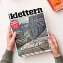 Schräg auf einem weißen Tisch liegende Zeitschrift „Klettern – Abenteuer | Lifestyle | Sport“, von zwei Händen gehalten, mit grauen Felswänden und einer kletternden Person im blauen Oberteil auf dem Titelbild. Sichtbare Texte: „ALPIN DE LUXE“, „HOTSPOT IM HIMALAYA“, „Kletterschuhe im großen Test“, „KÖRPER-SPANNUNG“ und „TESTSIEGER: Vom Klettergurt bis zum Schlafsack – die Produkt-Highlights 2026“; oben rechts liegt ein orangefarbenes Objekt.