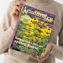 Schräg gehaltene Zeitschrift „Kraut&rüben“ mit Titelbild gelber Blüten und violettem Hintergrund, in den Händen einer Person mit beigem Strickpullover. Lesbare Texte: „Schöner. Wilder. Biogarten.“, „Cool-Flower-Methode: Jetzt säen, im Frühling strahlen“, „Gärtnern im Schein der späten Sonne“ und „Die Tomaten sind reif!“.