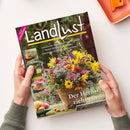 Schräg gehaltene Zeitschrift „Landlust“ mit schwarzem Cover, großem weißem Titel und pinkem Hinweis „NEUE STRICKIDEEN“. Abgebildet sind ein üppiger herbstlicher Blumenstrauß in einem Topf, mehrere kleine Garten- und Bastelfotos sowie die Texte „Laubsammler: Aufsatz für Schubkarren bauen“, „Basteln: Blüten aus Kürbiskernen“, „Unverzichtbar: Ehrenamtliche Bürgermeister“ und „Der Herbst zieht ein“; zwei Hände mit grünen Strickbündchen halten das Heft, das Geschlecht ist nicht erkennbar.