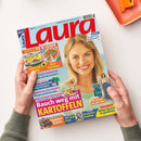 Auf weißem Untergrund halten zwei Hände ein schräg liegendes, glänzendes Zeitschriftencover der „Laura“ mit rotem Titel, blauem Hintergrund und einer blondhaarigen weiblichen Person. Sichtbare Texte: „BEST SELLER“, „IMMER FIX & LECKER“, „Ist vegan essen wirklich gesund?“, „Bauch weg mit Kartoffeln“, „Neue Pizza-Ideen“, „Warnsignale früher erkennen“, „Die perfekte Haarpflege“ und „Urlaubs-Glück“.