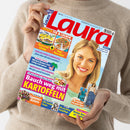 Frau im beige gestrickten Pullover hält eine schräg fotografierte, farbig gestaltete „Laura“-Zeitschrift mit hellblauem Cover und großem rotem Titel. Sichtbare Texte: „IMMER FIX & LECKER“, „Kuchen mit Schwips“, „Neue Pizza-Ideen“, „Mein Sohn will nicht ausziehen“, „Günstig reisen“, „Allergien gezielt behandeln“, „Bauch weg mit Kartoffeln“, „Sommer-Astern“ und „Die perfekte Haarpflege“; auf dem Cover sind ihr Gesicht, Speisen, Blumen und weitere Fotomotive abgebildet.