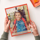 Rot gerahmtes Elternmagazin „leben & erziehen“, schräg von oben fotografiert und von zwei Händen gehalten. Auf dem Cover lächelt ein Mädchen mit braunem Dutt in Jeanslatzhose, gestreiftem Shirt und rotem Rucksack; lesbare Texte sind „DAS ELTERN-MAGAZIN“, „REISE NACH MAROKKO“, „FAMILIEN-FRIEDEN“, „SELBST-FÜRSORGE“, „AUFS BAUCHGEFÜHL HÖREN“ und „Hallo Schule“. Oben rechts liegt ein orangefarbenes Heftgerät auf weißer Fläche.