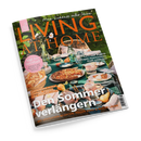 Schräg liegendes Magazin „LIVING AT HOME“ mit grünem Gartenmotiv und gedecktem Tisch: Geschirr, Gläser, Tassen, Blumen, Obst und Kuchen auf einer türkisfarbenen Tischdecke, dahinter ein Korbstuhl. Sichtbare Texte: „Mein Zuhause voller Ideen“, „Süß bis herzhaft“, „Citytrip Porto“, „Spätsommer-Glück“ und „Der Sommer verändert“.