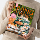 Weibliche Person im beigefarbenen Strickpullover hält eine schräg ausgerichtete Zeitschrift mit dem Titel „LIVING AT HOME“ und dem Text „Mein Zuhause voller Ideen“. Das Cover zeigt einen gedeckten Tisch mit Speisen, Geschirr und Blumen sowie die Aufschriften „Gute Laune in der Küche“, „Citytrip Porto“, „Den Sommer verlängern“ und „Mit köstlichen Rezepten, toller Deko & bester Laune“.