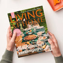 Schräg gehaltenes Magazin „LIVING AT HOME“ mit dem Titel „Mein Zuhause voller Ideen“ und Garten-Tischszene mit Kuchen, Geschirr, Gläsern, Blumen und Getränken. Sichtbare Texte: „Gute Laune in der Küche“, „Citytrip Porto“, „Der Sommer verlängern“ sowie „Spätsommer-Glück: Dahlien, Zinnien und Sonnenblumen“; zwei Hände halten das Magazin.