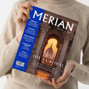 Weibliche Person im cremefarbenen Strickpullover hält ein schräg abgebildetes MERIAN-Reisemagazin mit blauem Rand und braunem Titelbild. Lesbare Texte: „MERIAN“, „The Art of Travel“, „OH EUROPA!“, „Albanien“, „Finnland“, „Starke Städte“, „Lüneburger Heide“ und „Istrien“; das Titelbild zeigt ein steinernes Tor mit Blick auf eine Kuppel im warmen Abendlicht.