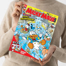 Weibliche Person im beigefarbenen Strickpullover hält ein schräg ausgerichtetes „Micky Maus“-Magazin mit gelbem Titelbereich und roter Schrift „Micky Maus“ sowie „Disney“-Logo. Das Cover zeigt Donald Duck und weitere Entenfiguren, umgeben von fliegenden Münzen und blauen Explosionseffekten; lesbare Texte: „Top-Comic: Geniale Geldwäsche“, „W.I.T.T.“ und „Spiegel-Trick“.