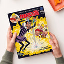 Schräg von oben fotografiertes, glänzend bedrucktes „mosaik“-Comicheft in zwei Händen mit grauen Ärmeln vor weißem Hintergrund. Das Cover zeigt mehrere gezeichnete Figuren, einen Zauberer mit erhobenen Armen, gelbe Explosions- und Rauchdarstellungen sowie die Texte „ABENTEUER IN DER GOLDENEN STADT“, „DIE JAGD NACH DEM GOLDE“ und „Empfohlen von Stiftung Lesen“; oben rechts liegt ein orangefarbenes Tablett.