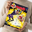Hochformatiges „mosaik“-Comicheft aus glänzendem Papier, schräg von oben fotografiert und von einer Person ohne sichtbares Gesicht im beigen Strickpullover gehalten. Dunkelblaues Cover mit gelber Explosion, mehreren gezeichneten Figuren und den Texten „ABENTEUER IN DER GOLDENEN STADT“, „DIE JAGD NACH DEM GOLDE“ sowie „Empfohlen von Stiftung Lesen“.