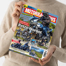 Eine Person im beigefarbenen Strickpullover hält ein schräg ausgerichtetes Motorradmagazin mit blau-rotem Cover. Sichtbare Texte: „MOTORRAD“, „Service: Motorrad und Hitze“, „Retro-Hornet“, „Fahrbericht: Honda CB 1000 F“, „Scrambler-Vergleich Ducati vs. Triumph“, „Gebrauchtberatung Kawasaki W800“, „Yamaha XT 500“, „Reisefieber“ sowie „Schottland & Türkei“.