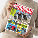 Weibliche Person im beigefarbenen Strickpullover hält eine schräg fotografierte Zeitschrift mit dem Titel „MOTORRAD“ und der Aufschrift „GRÖSSTE MOTORRADZEITSCHRIFT“; das Cover zeigt mehrere Motorräder. Sichtbare Themen: „Dauertest KTM 1390 Super Duke R Evo“, „Traum-Touren strecken in Kärnten“, „MotoGP Interview mit Marco Bezzecchi“, „Japan … Top-Tester“, „BMW-Neuheiten“ und „Gebrauchtkauf Ducati Panigale V4/V4 S“.
