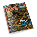Schräg freigestelltes Magazin „mountainBIKE“ mit orangefarbener Klebebindung und glänzendem Cover vor schwarzem Hintergrund; abgebildet sind ein bewachsener Mountainbike-Trail, eine radfahrende Person sowie Fahrräder auf einem Heckträger neben einem Auto. Sichtbare Texte: „E-MTB“, „ALLES IST FAHRBAR“, „Kupplungsträger“, „14 XC-Reifen im Labor- & Praxistest“, „BOCK auf SPEED“, „9 WM-Bikes im Test“ und „E-CVT“.