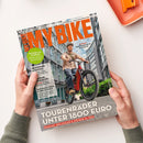 Aufgeschlagenes Magazin „BIKE“ mit orangefarbener Titelgestaltung, gehalten von zwei Händen; auf dem Cover fährt eine männliche Person mit Helm ein rotes Fahrrad zwischen Hochhäusern. Sichtbare Texte: „RAD & E-BIKE · TECHNIK · REISEN“, „Blinker am Fahrrad – Praxis-Check & Rechtslage“, „Brandneu Specialized Vado 3X“, „Panorama-Tour in der Schweiz“ und „Diamant, Decathlon & Stevens im Test“.
