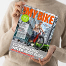 Hände einer Person in beigefarbenem Strickpullover halten schräg ein Fahrradmagazin mit orangefarbenem Titel „BIKE“. Das Cover zeigt einen männlichen Radfahrer mit Helm auf einem roten Fahrrad vor modernen Gebäuden; sichtbar sind außerdem die Aufschriften „NEWCOMER AVENTON“, „FAHRRAD-RUCKSÄCKE“, „PANORAMA-TOUR IN DER SCHWEIZ“ und „TOUREN-RADLER UNTER 1800 EURO“.