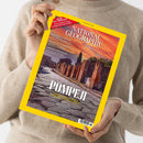Eine Person ohne sichtbares Gesicht hält ein schräg ausgerichtetes Magazin mit gelbem Rand über einem beigefarbenen Strickpullover. Auf dem Cover stehen „NATIONAL GEOGRAPHIC“, „AUGUST 2026“ und „POMPEJI – LEBEN UND STERBEN IM SCHATTEN DES VESUV“; abgebildet sind antike Ruinen unter einem orangefarbenen Himmel.