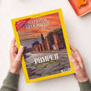 National-Geographic-Magazin mit gelbem Rand, dem Titel „NATIONAL GEOGRAPHIC“ und der Datumsangabe „AUGUST 2026“, aufgeschlagen von zwei Händen über einem weißen Tisch. Das Cover zeigt Ruinen in Pompeji bei orangefarbenem Himmel sowie die Texte „POMPEJI“, „LEBEN UND STERBEN IM SCHATTEN DES VESUV“ und weitere Themenhinweise.
