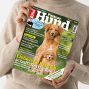 Person im beigefarbenen Strickpullover hält ein schräg fotografiertes Magazin mit grünem Cover, dem Titel „Partner Hund“ und Bildern eines ausgewachsenen roten Hundes sowie eines Welpen. Sichtbare Texte: „Liebevolle Klarheit schafft Vertrauen“, „Echte Expertenwissen: Kleine Hunde erziehen“, „Fakten-Check Nass- oder Trockenfutter“ und „Unsere Rasse-Porträts“.