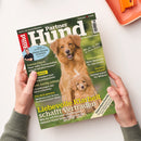 Schräg von oben fotografierte Zeitschrift „Partner Hund“ auf weißer Fläche, von zwei Händen einer nicht näher erkennbaren Person gehalten. Das grüne Cover zeigt einen braunen Hund und einen Welpen sowie die Texte „Kleine Hunde erziehen“, „Osteopathie“, „Mehr Mobilität für den Senior“ und „Liebevolle Klarheit, liebevolle Vertrauensschaft“; oben rechts liegt ein orangefarbener Gegenstand.