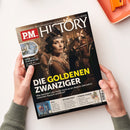 Aufgeschlagenes Magazin „PM-HISTORY“ in zwei Händen über weißer Fläche, mit orangefarbenem Notizbuch und Klammer im Hintergrund. Das Titelbild zeigt eine weibliche Person im historischen Kostüm mit Feder und die Texte „Europas größtes Monatsmagazin für Geschichte“, „DIE GOLDENEN ZWANZIGER“, „MEHR ALS NUR MUSTER“, „9/11“, „BUCHKUNST“ und „TOMOE GOZEN“.