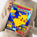 Hochformatiges, glänzendes Pokémon-Magazin, diagonal von einer Person in beigefarbenem Strickpullover gehalten. Das Cover zeigt einen großen gelben Pikachu vor blauem Hintergrund mit Konfetti sowie die Texte „POKÉMON“, „Schnapp sie dir alle!“, „HIGHLIGHT-AUSGABE“, „3D-ANHÄNGER“, „FUNPACK“, „XL-POSTER“, „POSTKARTEN“, „Mit Mega-Gewinnspiel!“ und „4 Postkarten mit Goldprägung!“.