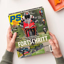 Aufgeschlagenes, grün gestaltetes Motorradmagazin „PS Sport motorrad magazin“, von zwei Händen mit grünen Strickärmeln gehalten; darauf eine rote Sportmaschine mit Motorradfahrperson sowie mehrere kleinere Motorräder. Sichtbare Texte: „Zonkos Erfahrungen: Wie ich Motorradjournalist wurde“, „Die neue Mittelklasse – besser als legendäre Superbikes?“, „Feine Zweitakt-Renner“, „Rekord Supertest“ und „BMW M 1000 RR“; oben rechts liegt ein orangefarbenes Objekt.