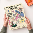 Aufgeschlagene, cremefarbene Zeitschrift „PSYCHOLOGIE HEUTE“ mit illustrierten weiblichen Personen und farbigen Formen auf dem Titel, von zwei Händen gehalten. Sichtbare Texte: „Konflikte leichter lösen“, „ADHS und Autismus – Ein Leben zwischen zwei Extremen“, „Abnehmen – Verändert es die Liebe?“ und „Arbeitsweg – Kreative Lösungen gegen Stress“; daneben liegt ein orangefarbenes Notizbuch mit Büroklammer.
