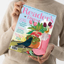 Person in beigefarbenem Strickpullover hält ein schräg abgebildetes Magazin mit hellblauem Cover. Der Titel „Reader’s Digest“ sowie „HEALTH • MONEY • TRAVEL • FOOD & DRINK • CULTURE • REAL STORIES“ sind sichtbar; darunter stehen „6 Easy Ways To Get A Greener Thumb“ und „Sean Penn On Politics, Pressure And Perseverance“. Das Cover zeigt eine farbige Blumenillustration mit zwei Vögeln, Blättern, Beeren und einer Schnecke.